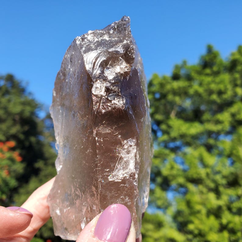 May include: A large, translucent smoky quartz crystal with a rough, natural texture. The crystal is a light brown color and is held against a backdrop of a blue sky and green foliage.