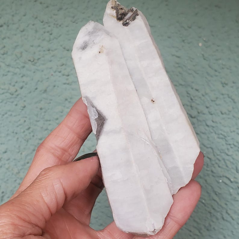 Pu&ograve; includere: Un gruppo di due cristalli di quarzo bianco dall'aspetto leggermente traslucido. I cristalli hanno una consistenza naturale e ruvida e sono tenuti in mano su uno sfondo verde chiaro. Alcune aree presentano inclusioni grigio scuro.