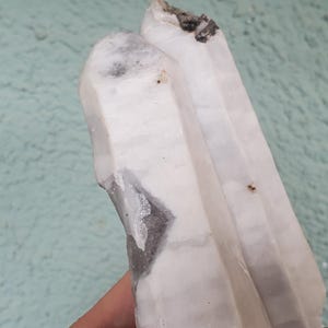 Pu&ograve; includere: Primo piano di un gruppo di cristalli bianchi con una superficie ruvida e strutturata. Il cristallo presenta aree di inclusioni grigie e nere. Lo sfondo &egrave; di colore verde acqua chiaro.
