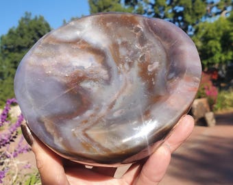 Puede incluir: Una piedra pulida de forma ovalada con patrones arremolinados de color marrón, blanco y lavanda. La piedra se sostiene en una mano, con un fondo natural al aire libre de árboles y cielo azul. La superficie de la piedra refleja la luz.
