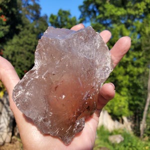 May include: A large, translucent smoky quartz crystal held in an open hand. The crystal has a rough, irregular shape with a light brown hue and visible internal structures. The background shows green foliage and a blue sky.