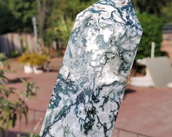 May include: A polished moss agate crystal tower featuring a pointed top and a cylindrical body. The stone displays a translucent white base with intricate, branching dark green inclusions that resemble moss or fern patterns throughout the surface.