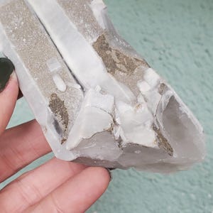 Pu&ograve; includere: Un gruppo di cristalli di quarzo grezzo e naturale. Il cristallo presenta un mix di sezioni bianche chiare e opache, con inclusioni beige e grigie. La consistenza ruvida e la forma irregolare ne evidenziano l'origine naturale. Un esemplare minerale unico.