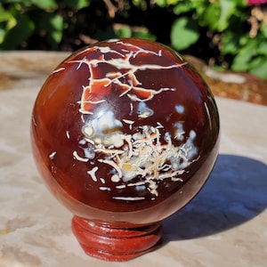May include: A polished, spherical gemstone with a rich, dark reddish-brown hue, accented by intricate white and cream-colored veining. The sphere sits on a small, round, reddish-brown wooden stand, showcasing its natural beauty.