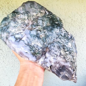 May include: A large, raw piece of moss agate featuring a translucent, milky white base with deep green, moss-like inclusions and dark, jagged mineral veins throughout the stone. The surface has a polished, glossy texture with irregular edges.