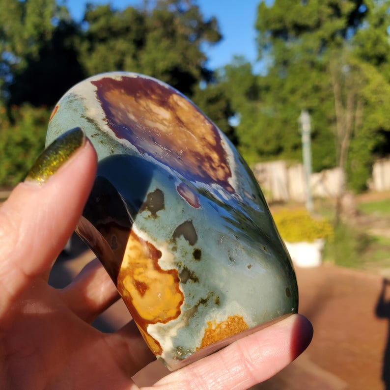 May include: A polished, irregularly shaped stone with a mix of colors. The stone features areas of brown, tan, and teal, with a glossy finish. The stone is held in a hand, with a blurred background of trees and sunlight.