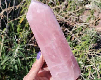 May include: A polished rose quartz crystal tower with a hexagonal shape and a pointed top. The stone features a translucent, pale pink color with natural internal fractures and variations in opacity throughout the faceted surface of the crystal.