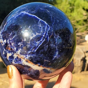 Könnte beinhalten: Ein polierter, kugelförmiger Sodalith-Stein mit tiefblauen Farbtönen, komplexer weißer Calcit-Aderung und subtilen orangefarbenen Mineraleinschlüssen. Die Oberfläche ist glatt und reflektierend und zeigt ein natürliches, marmoriertes Muster.