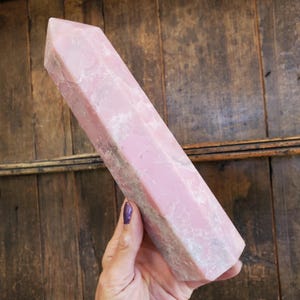 May include: A large, pink crystal obelisk with a faceted top. The stone has a marbled appearance with white and gray veining. The obelisk is held in a hand against a wooden background.
