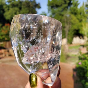 Puede incluir: Un cristal claro y facetado con fracturas e inclusiones internas. El cristal se sostiene contra un fondo verde borroso. La superficie del cristal refleja la luz, creando un efecto brillante. El cristal es una gema natural y en bruto.