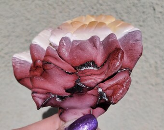 Puede incluir: Escultura decorativa de una flor, hecha a mano. Presenta un diseño en capas con tonos rojos, blancos y beige. Detalles en negro acentúan los pétalos. La escultura es sostenida por una persona con esmalte de uñas morado.