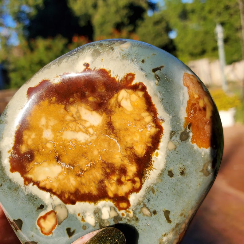 May include: A polished stone with a rounded shape. The stone features a mix of colors, including a base of pale blue-green, with large patches of brown and yellow-beige. The stone has a smooth, glossy surface and a natural, organic appearance.
