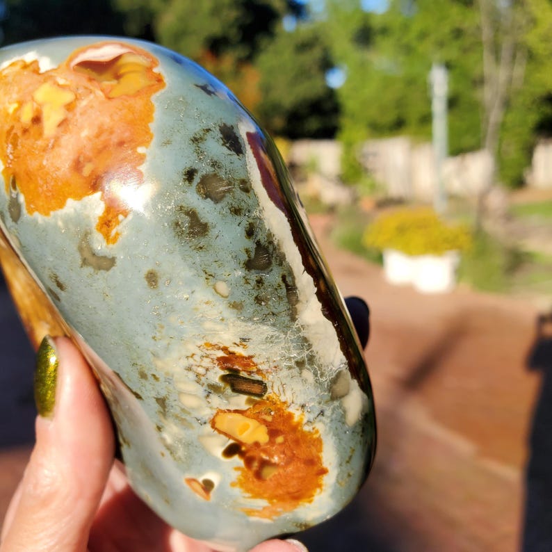 May include: A polished, oblong stone with a mix of colors. The stone features a base of pale blue-green with patches of orange-brown and yellow. The surface has a glossy finish, and the stone is held in a hand.