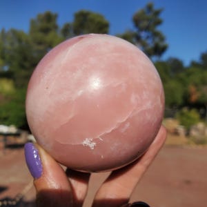 Peut inclure: Une grande sph&egrave;re rose lisse, possiblement une boule de cristal ou une pierre d&eacute;corative. La sph&egrave;re pr&eacute;sente de subtiles veines blanches et une surface l&eacute;g&egrave;rement irr&eacute;guli&egrave;re. L'arri&egrave;re-plan pr&eacute;sente des arbres verts et un ciel bleu.