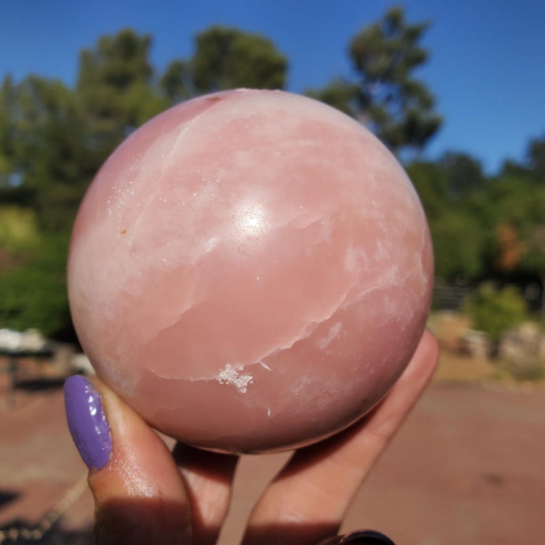 Peut inclure: Une grande sph&egrave;re rose lisse, possiblement une boule de cristal ou une pierre d&eacute;corative. La sph&egrave;re pr&eacute;sente de subtiles veines blanches et une surface l&eacute;g&egrave;rement irr&eacute;guli&egrave;re. L'arri&egrave;re-plan pr&eacute;sente des arbres verts et un ciel bleu.