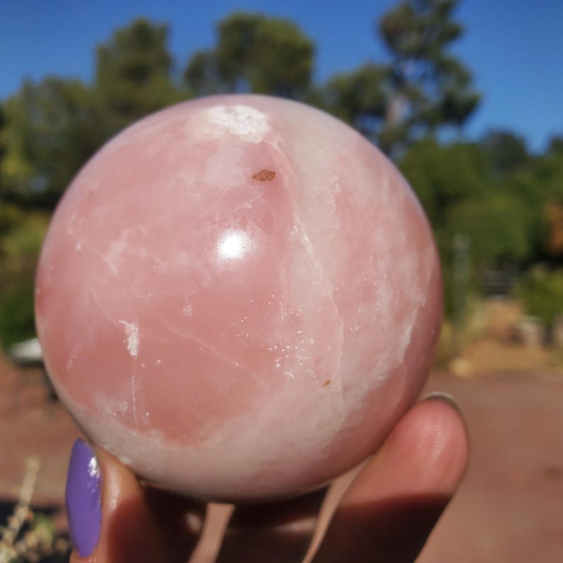 Peut inclure: Une boule de cristal en quartz rose polie. La sph&egrave;re est d'un rose tendre avec des veines blanches et quelques petites inclusions brunes. L'arri&egrave;re-plan est flou, avec des arbres verts et un ciel bleu.