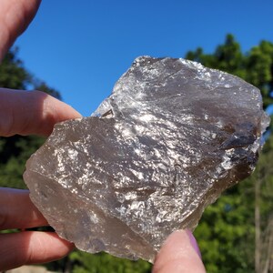 May include: A rough, translucent smoky quartz crystal with a natural, irregular shape. The crystal has a light brown color and reflects light, creating a shimmering effect. The background is a bright blue sky and green foliage.