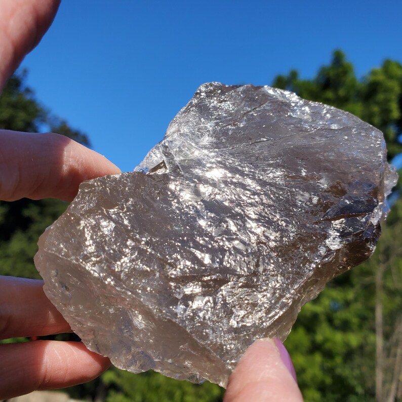 May include: A rough, translucent smoky quartz crystal with a natural, irregular shape. The crystal has a light brown color and reflects light, creating a shimmering effect. The background is a bright blue sky and green foliage.