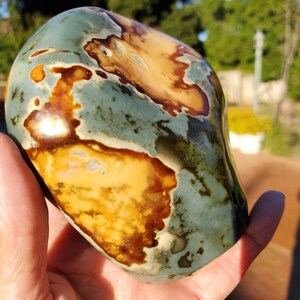 May include: A polished stone with a smooth, rounded shape. The stone features a blend of colors, including teal, brown, and yellow, creating a unique pattern. The stone is held in a hand, showcasing its size and texture.