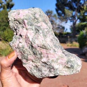 May include: A raw, unpolished mineral specimen featuring a rough, jagged texture with a mottled appearance. The stone displays a combination of opaque, light pink crystalline sections interspersed with dark, forest green mineral inclusions.