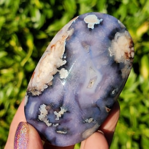 Könnte beinhalten: Ein polierter, ovaler Achatstein mit einer durchscheinenden violetten und lavendelfarbenen Basis, komplexen weißen, wolkenartigen Einschlüssen und kleinen Flecken aus rotbraunen Mineralablagerungen auf seiner glatten, glänzenden Oberfläche.