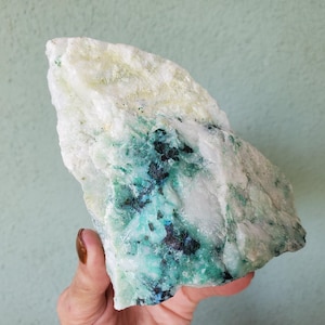 May include: A raw mineral specimen featuring a combination of opaque white quartz, vibrant green inclusions, and dark black crystalline patches. The stone has an irregular, jagged shape with a rough, textured surface and varied mineral deposits.