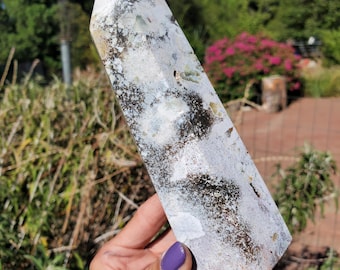 May include: A polished, hexagonal crystal tower featuring a mottled pattern of white, gray, and translucent pale green mineral inclusions, accented by dark brown to black dendritic or moss-like organic-looking patches throughout the stone.