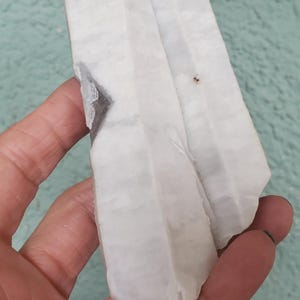 Pu&ograve; includere: Un cristallo bianco grezzo con un'inclusione grigio scuro. Il cristallo &egrave; tenuto in mano, mostrando la sua consistenza naturale e ruvida. Lo sfondo &egrave; di colore verde acqua chiaro.