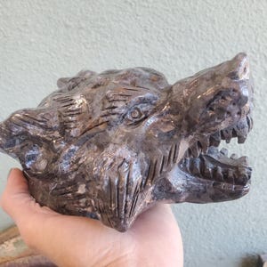 Puede incluir: Una escultura detallada de la cabeza de un lobo, tallada en piedra oscura y moteada. La boca del lobo está abierta, revelando afilados dientes, y su pelaje está intrincadamente detallado con líneas talladas. La escultura se sostiene en una mano.