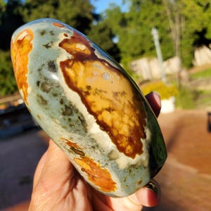May include: A polished, oval-shaped stone with a blend of colors. The stone features areas of orange, brown, yellow, and blue-green hues. The surface has a smooth, glossy finish, highlighting the natural patterns and variations within the stone.