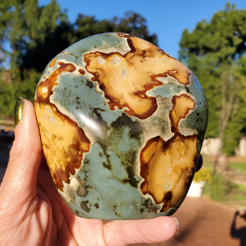 May include: A polished, rounded stone with a unique pattern resembling a map. The stone features a blend of colors, including tan, brown, and blue-green hues. The smooth surface reflects light, highlighting the intricate details of the natural design.