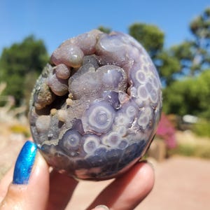 May include: A close-up of a geode with a mix of purple and gray hues. The geode has a bubbly, textured surface with circular patterns. The background is blurred, showing greenery and a bright blue sky.
