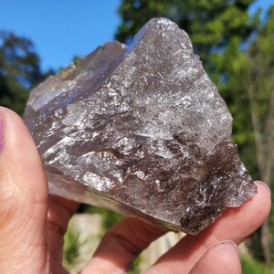 May include: A large, rough-cut smoky quartz crystal with a translucent appearance. The crystal has a dark, smoky brown color with areas of lighter, clear quartz. The surface reflects light, creating a sparkling effect. The crystal is held in a hand.