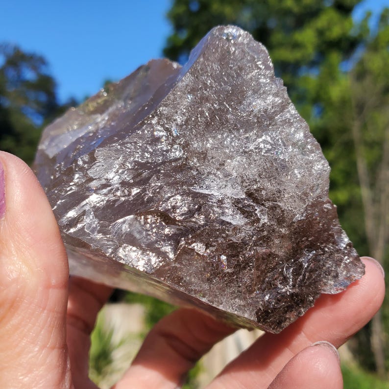 May include: A large, rough-cut smoky quartz crystal with a translucent appearance. The crystal has a dark, smoky brown color with areas of lighter, clear quartz. The surface reflects light, creating a sparkling effect. The crystal is held in a hand.