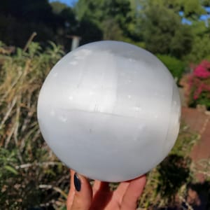 Puede incluir: Una bola de cristal de selenita esférica y lisa con un aspecto blanco lechoso y translúcido. La superficie muestra una textura fibrosa sutil y un suave brillo nacarado que capta la luz, creando un efecto resplandeciente en la forma redonda.