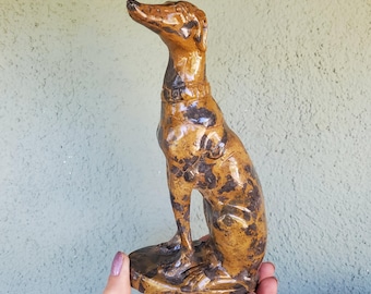 Puede incluir: Una figura de perro de cerámica en posición sentada, con un acabado brillante y un patrón moteado de colores marrón oscuro y tostado. El perro tiene un collar esculpido alrededor del cuello y está montado sobre una base circular, vista de perfil.