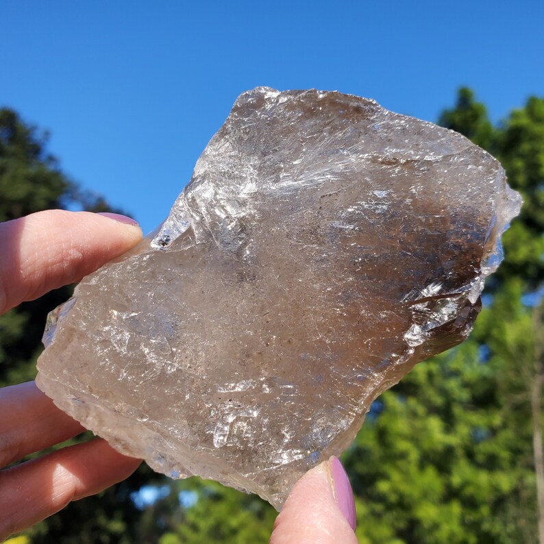 May include: A translucent, smoky quartz crystal with a rough, irregular shape. The crystal is held up against a backdrop of a bright blue sky and green foliage. The stone has a natural, earthy appearance.