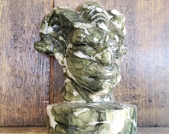 Puede incluir: Una escultura de piedra tallada de una cabeza de carnero con acabado pulido, que presenta un patrón veteado de color verde bosque profundo y blanco. El carnero se muestra con cuernos curvados y una estructura facial detallada sobre una base.