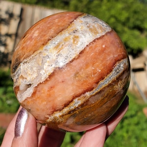 May include: A polished, spherical gemstone with bands of orange, white, and brown. The stone is held in a hand, showcasing its smooth, rounded form. The natural patterns create a unique, earthy aesthetic, perfect for display or collection.