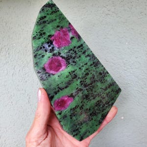 Könnte beinhalten: Ein polierter, unregelmäßig geformter Stein mit einer leuchtend grünen Basis, schwarzen Sprenkeln und auffälligen magenta-farbenen Rubin-Einschlüssen. Die Oberfläche des Steins ist glatt und reflektiert das Licht, wodurch seine natürliche Schönheit zur Geltung kommt.