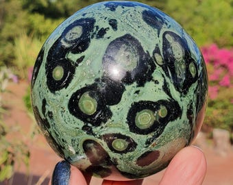 Puede incluir: Una piedra esférica pulida con un patrón complejo de marcas circulares en negro profundo y verde salvia. La superficie muestra un acabado liso y brillante con inclusiones orbiculares distintivas que crean un aspecto natural.