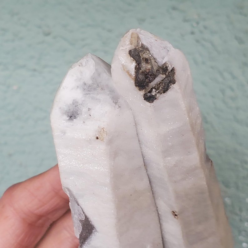 Pu&ograve; includere: Primo piano di un gruppo di cristalli di quarzo bianco con inclusioni grigio scuro e nere. Il cristallo ha una consistenza naturale e ruvida. Lo sfondo &egrave; di colore verde acqua.