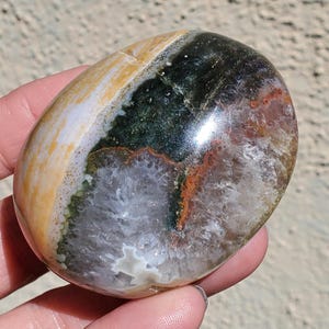 May include: A polished, oval-shaped gemstone with a mix of colors. The stone features bands of yellow, green, black, and red-brown hues. The center has a white, crystalline pattern. The stone is smooth and appears to be held in a hand.