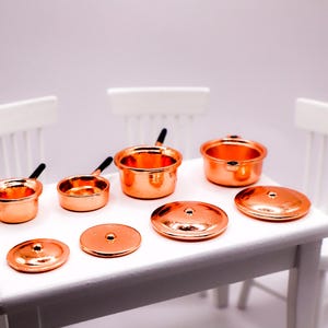 May include: Miniature copper cookware set including pots, pans, and lids. The set is arranged on a white table with white chairs in the background. The copper pots and pans have black handles.