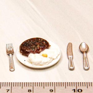 May include: Miniature dollhouse breakfast set. A small white plate holds a steak and fried egg. A fork, knife, and spoon are arranged around the plate. The set is about 2.5 cm wide.