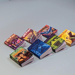 May include: A collection of miniature "Harry Potter" book replicas with colorful covers. The books are arranged on a light blue surface, showcasing various cover designs and titles from the series. The books are open, revealing the pages.
