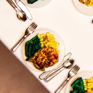 May include: Miniature plates of food with green beans, corn, and a meat dish, alongside tiny silverware. The plates are white, and the food is arranged to resemble a meal. The silverware includes forks, spoons, and knives.
