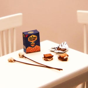 May include: Miniature s'mores scene featuring a box of Honey Maid graham crackers, two toasted marshmallows on sticks, two assembled s'mores, and a piece of foil-wrapped chocolate. The scene is set on a white surface.