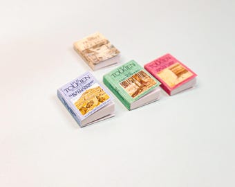 May include: Four miniature books with colorful covers are arranged on a white surface. The books have titles like "The Fellowship of the Ring" and "Two Towers" by Tolkien. The covers are in shades of blue, green, pink, and beige.