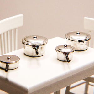 May include: Miniature silver pots and pans with lids sit on a white table, with two white chairs in the background. The pots have black handles. The scene is set in a miniature kitchen or dining room.